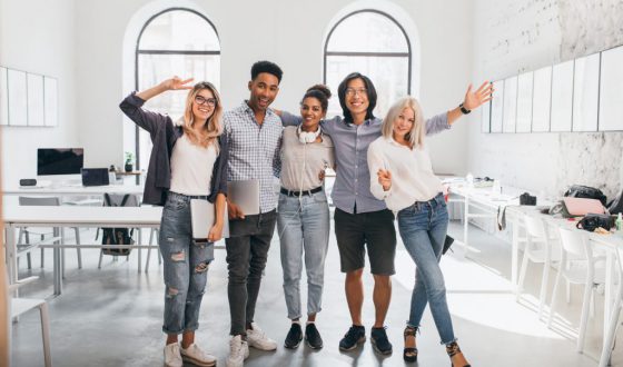 Full-length portrait of slim female office worker in jeans standing with legs crossed near asian colleague. Indoor photo of tall african student and glad european girl with laptop..