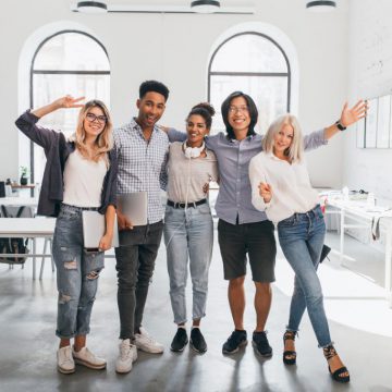 Full-length portrait of slim female office worker in jeans standing with legs crossed near asian colleague. Indoor photo of tall african student and glad european girl with laptop..